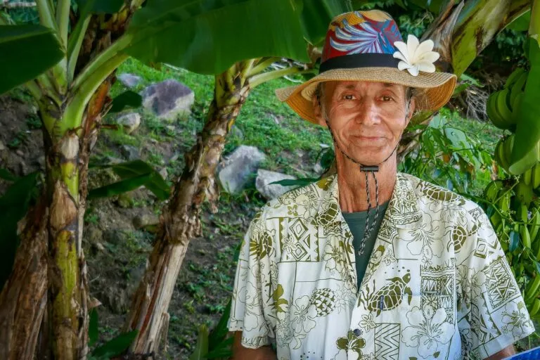 Smiling elderly man in a Hawaiian shirt and straw hat with a white flower, standing among banana plants in a tropical setting.