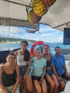 Six people sit closely on a boat, smiling for a group photo with the ocean and marina in the background, plus life rings and sky overhead.