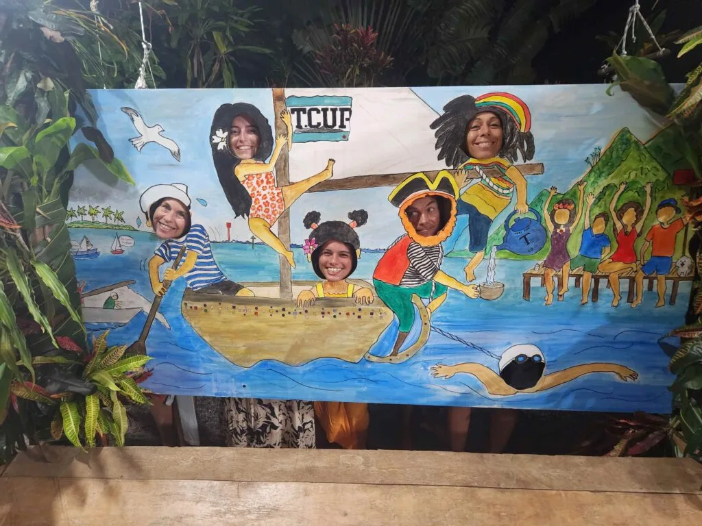 People Pose with Their Faces in Cut-out Holes of a Bright Mural Showing a Tropical Beach Scene with Boats and Cheering Crowd. 20260523_184659 20260523 184659 Régate La T cup Fête Ses 40 Ans
