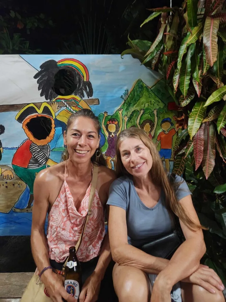 Two Smiling Women Pose in Front of a Colorful Mural Showing a Cheering Crowd and Rainbow, with a Beer Bottle in the Foreground. 20260523_184821 20260523 184821 Régate La T cup Fête Ses 40 Ans