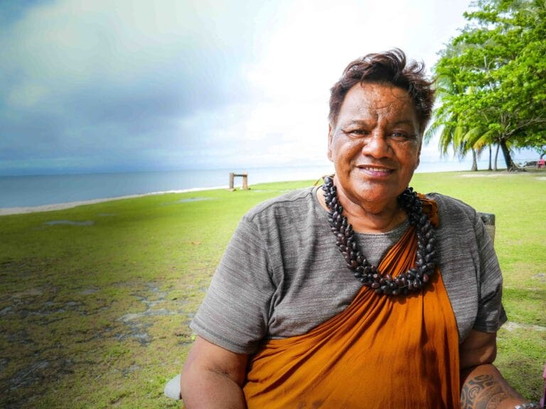 Close-up of a person wearing an orange robe and multiple dark bead necklaces, smiling outdoors by a grassy area and palm trees near the coast.