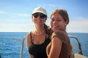Two friends posing for a boat selfie against a blue ocean and sky, one wearing a cap and sunglasses.