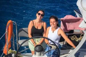 Two women aboard a sailboat smiling at the camera with the steering wheel and blue ocean behind them.