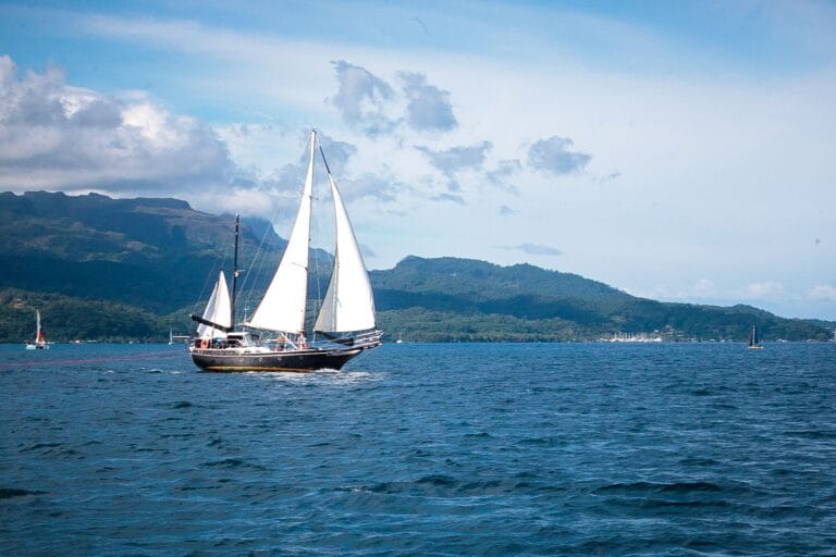 Sailing yacht with white sails on calm blue water, mountains in the distance under a partly cloudy sky
