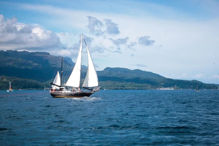 Sailing yacht with white sails on calm blue water, mountains in the distance under a partly cloudy sky