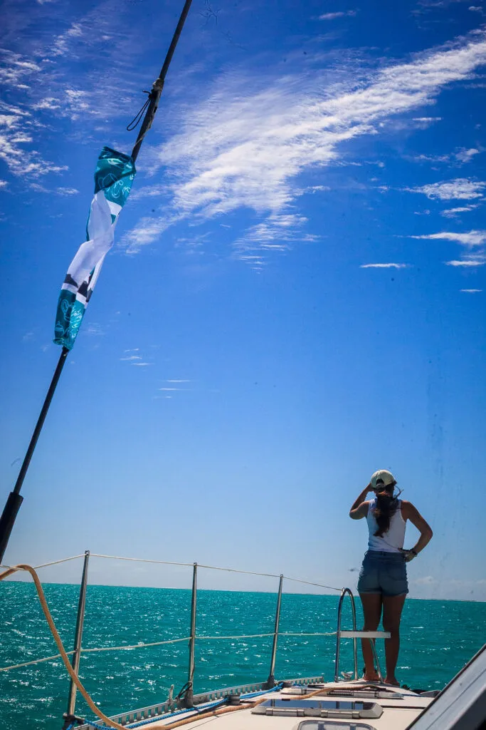 Person in a White Tank Top and Cap Stands on the Deck of a Sailboat, with a Teal Flag Flapping Against a Bright Blue Sky and Ocean. Tcupimg_3144-2026 Tcupimg 3144 2026 Régate La T cup Fête Ses 40 Ans