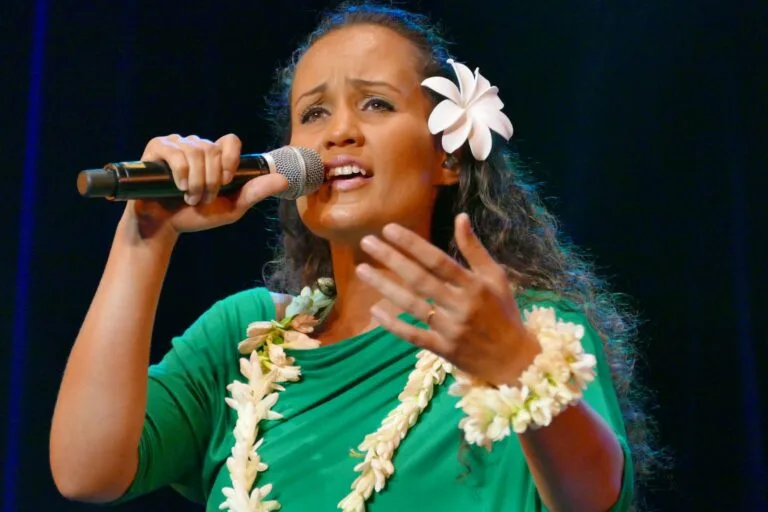 Female singer performing on stage, holding a microphone, wearing a green outfit and flower leis with a white hibiscus in her hair.